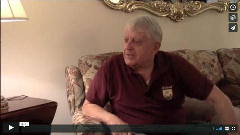 Screenshot from interview with Johnny Molloy, 2017 . Man wearing maroon shirt, seated on floral couch.