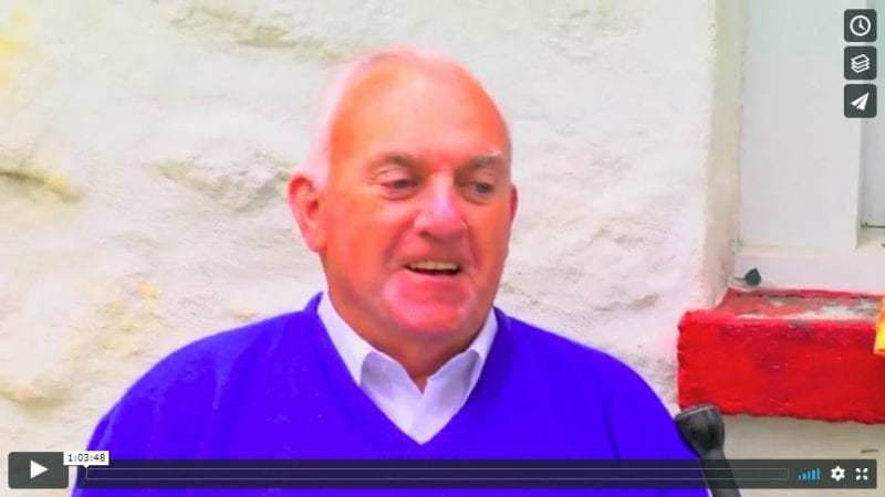 Screenshot from interview with Johnny Joyce, 2016. Head and shoulders image of man wearing purple sweater over white collared shirt.
