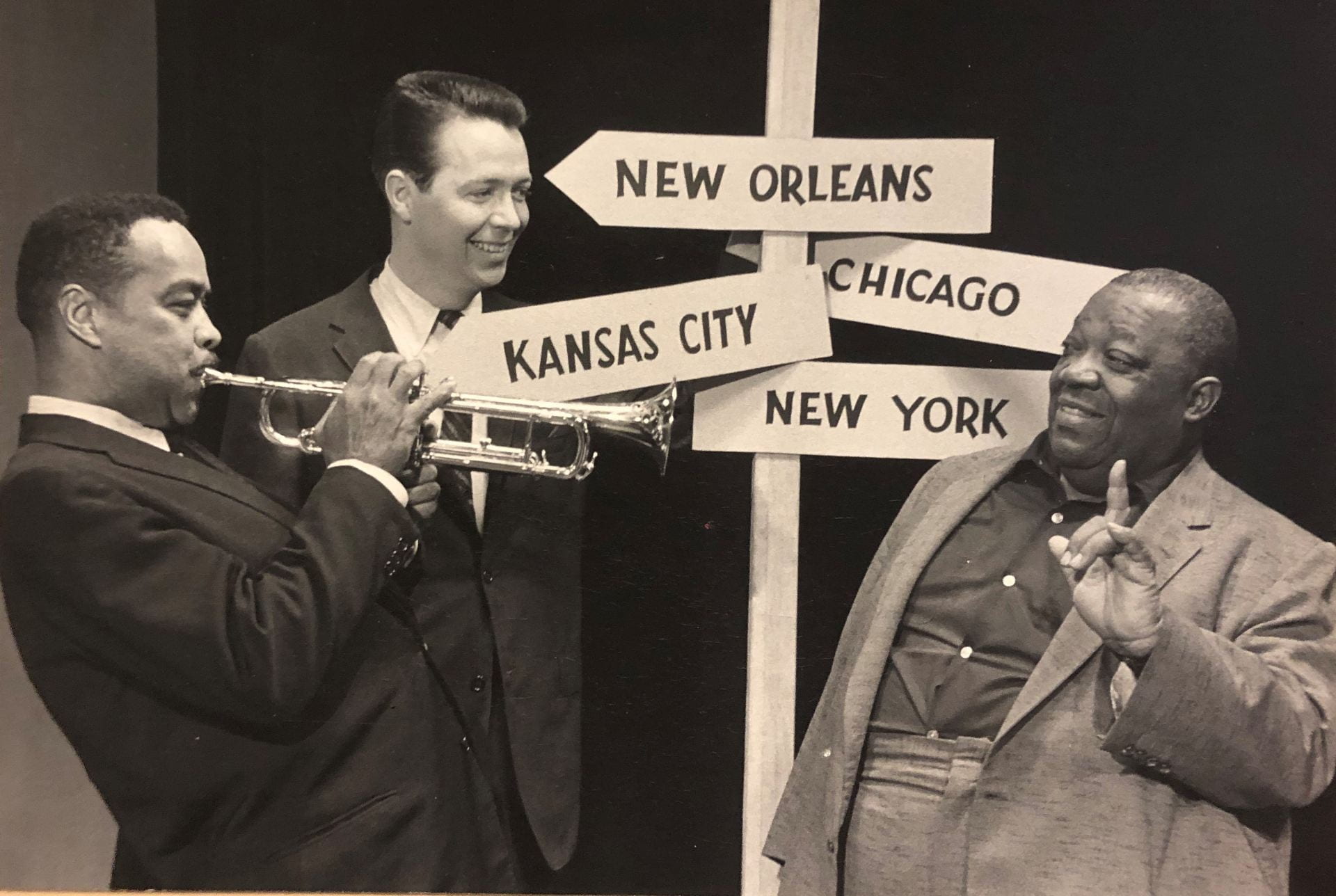 Black and white photograph from 1958 depicting radio announcer James Townsend Fitch standing with jazz musicians Clark Terry and Jimmy Ruching in front if a post with arrowed signs each labeled New Orleans, Kansas City, Chicago, and New York