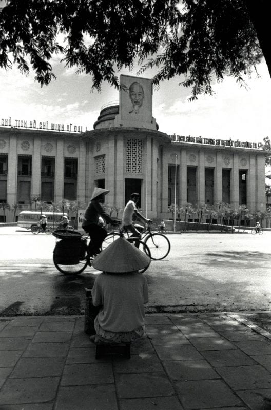 Woman sits on a sidewalk and people ride bicycles in front of the Central Bank