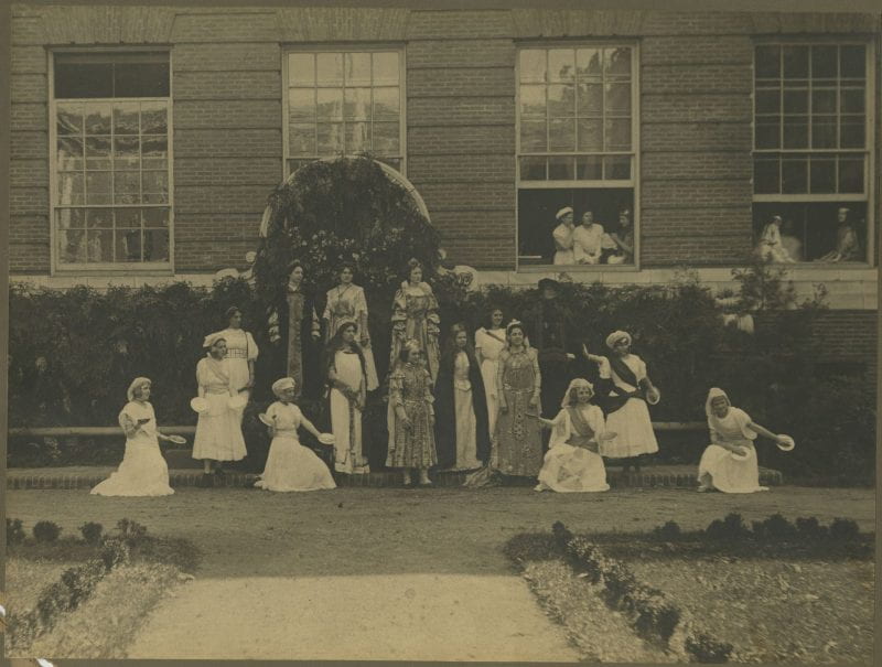 Black-and-white photo of young women performing a play outside of a school building