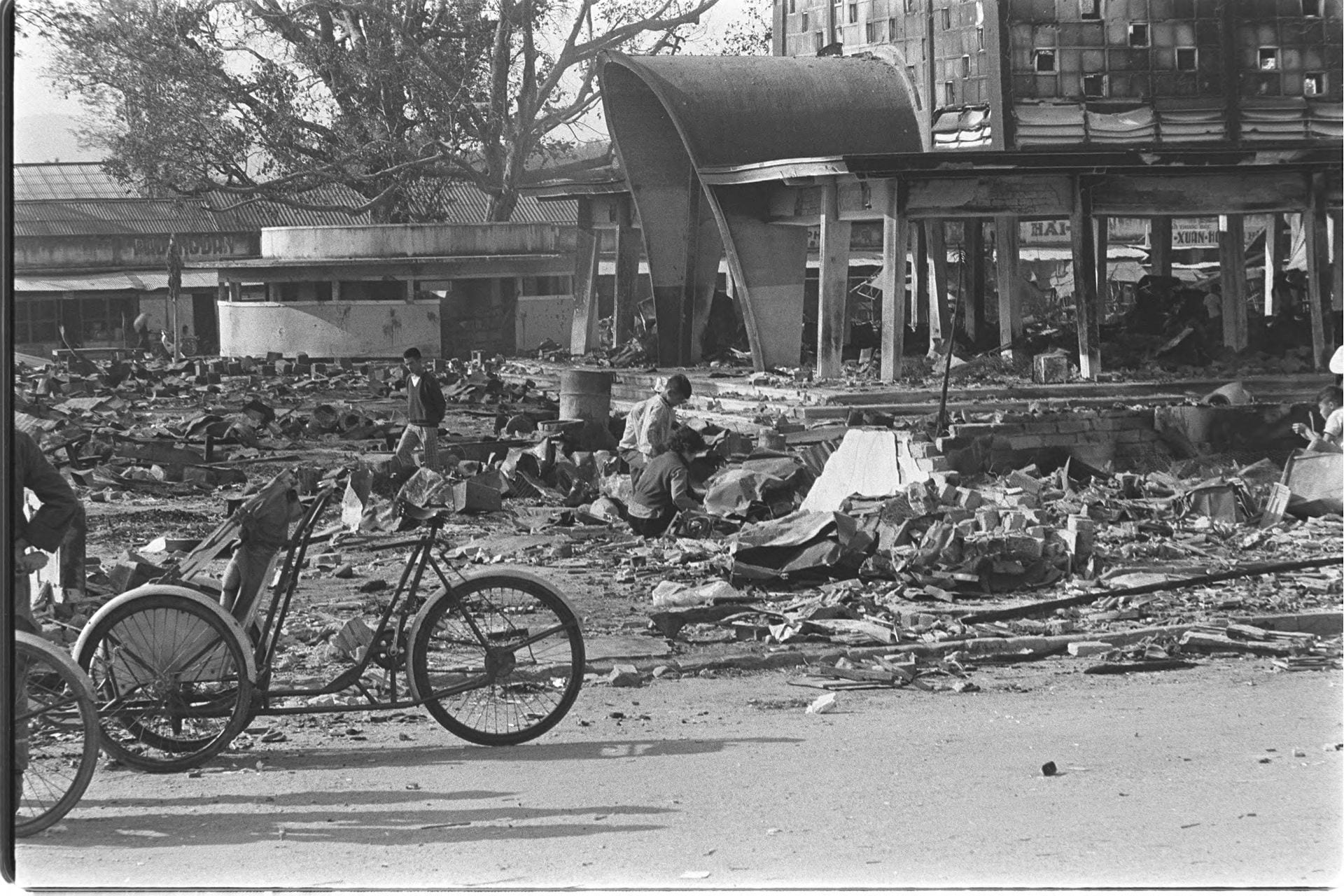 Black-and-white photo of people standing and crouching outside amongst building rubble and a bicycle in the foreground