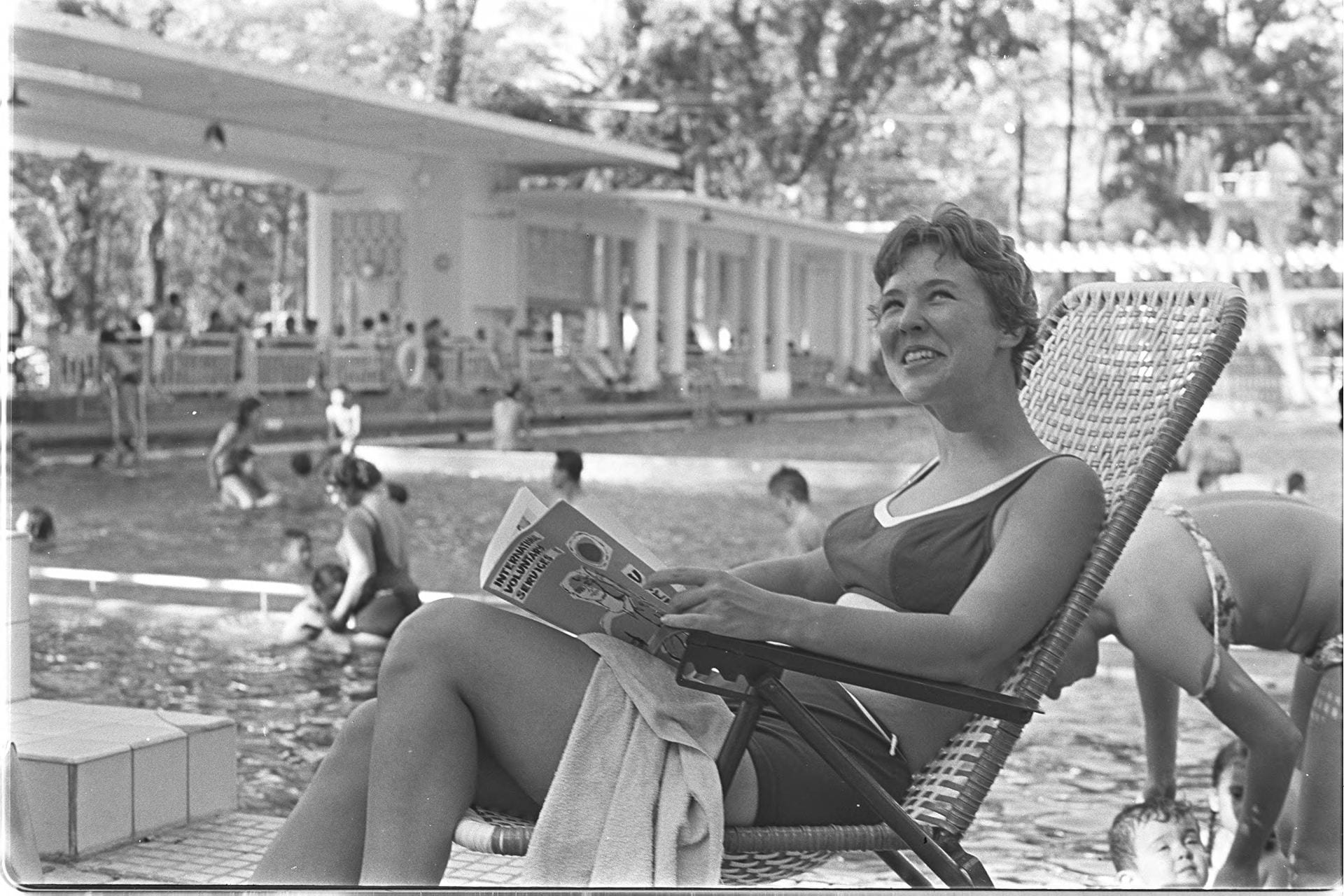 Black-and-white photo of a woman sitting in a pool chair with a magazine by a swimming pool