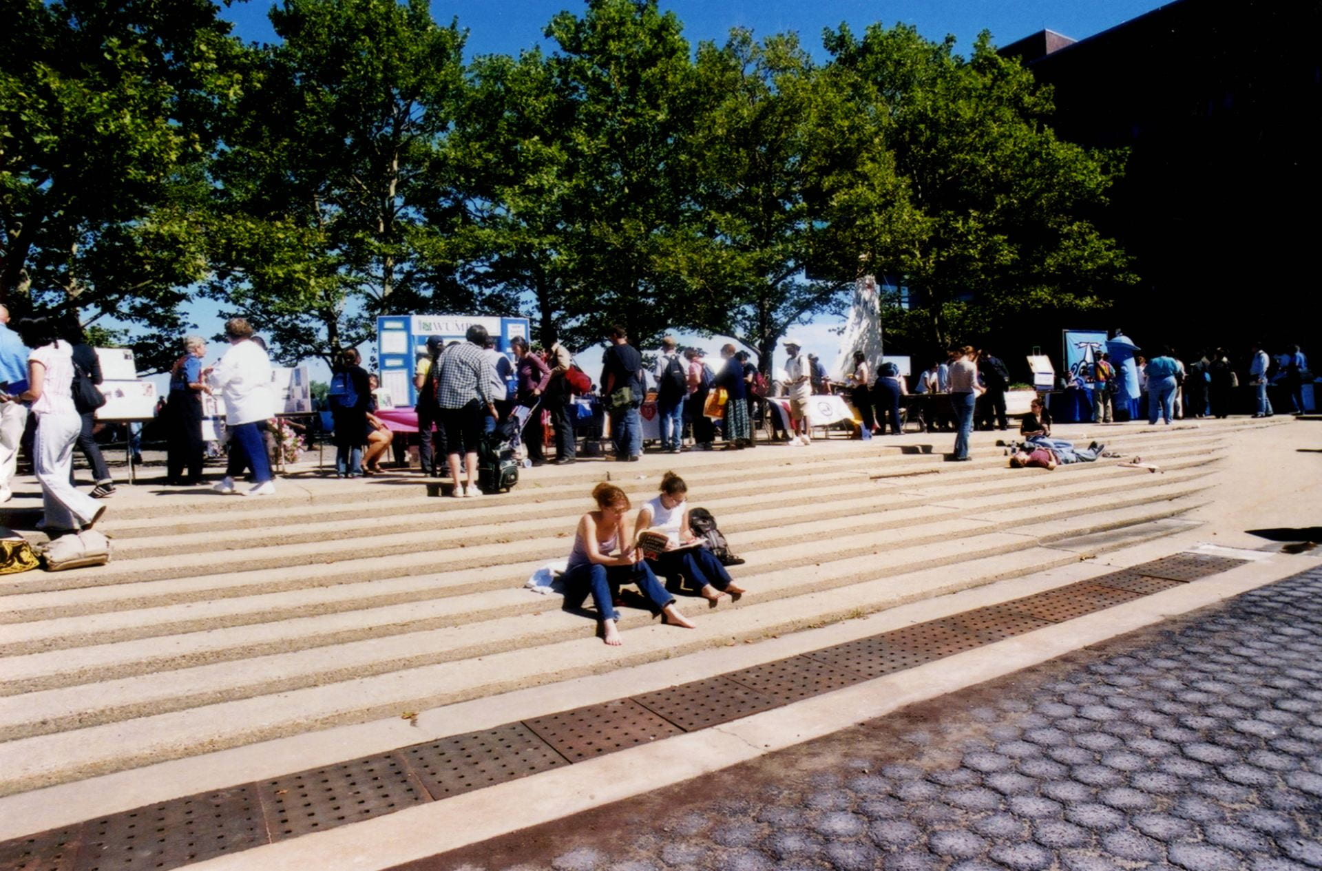 Color photograph of people looking at poster displays on the UMass Boston campus, with a few people sitting on steps in the foreground