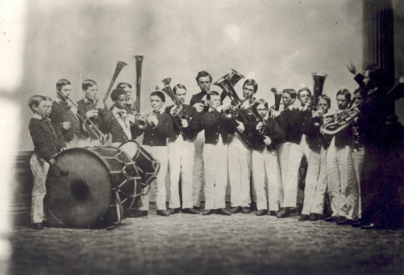A large group of young male students playing band instruments, including a bass drum, snare drum, cymbals, a trombone, a trumpet, French horns, tubas, and others. A conductor raising his arm stands in front of the group on the right.