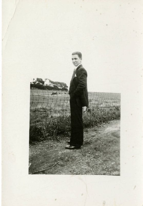 A young man stands on a dirt road facing a field with wire fencing at its fore edge. He is wearing a suit and dress shoes.