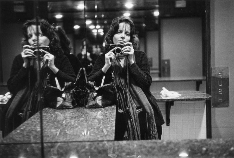 Woman stands in front of a mirror holding a camera, taking a picture of herself
