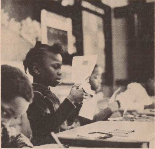 A young African American girl, likely 6-8 years old, sits in a classroom. She cuts a piece of paper carefully with scissors as a teacher helps another student on one side and a student cuts more paper on her other side.