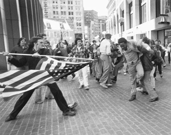 A white anti-desegregation protestor charges at a Black man with a sharp flagpole. 