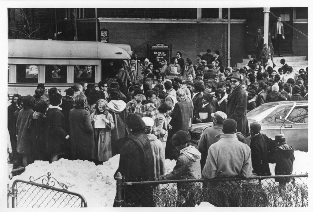 Photograph of a crowd of people standing outside a school.