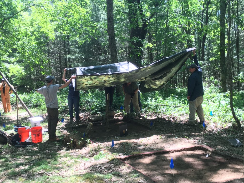 Archaeologists hold a tarp over an open excavation unit in order to take a photo