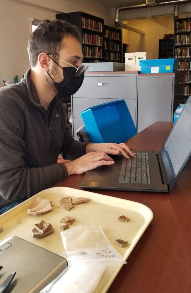 A student working at a computer with a tray of ceramic sherds.