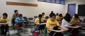 Students in yellow ALERTA t-shirt are focused and are sitting in the classroom with eyes glued on iPad. 