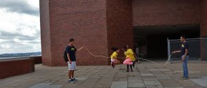 Two students jump rope together in the middle while mentors stand on the side holding up rope