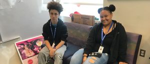 Two students sit on couch. Student on the left looks into camera while student on the right smiles.