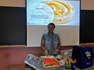 Behind is a photo of hummus. Infront of the screen, Daphney stands in front of aluminum container of carrots and celery with hummus and apple sauce on the side. Health and nutrition teacher stands in front of chalkboard ready to serve. 
