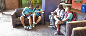 Two students sit on the couch on the left; One student looks over shoulder over another student as he plays game. Two students on the right also plays games on electronic device.