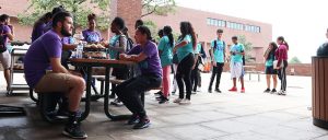 Students line up for lunch by picnic table where mentors in purple t-shirts sit.